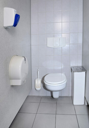 Ein helles und sauberes Badezimmer mit grauen Fliesen auf dem Boden und weißen Fliesen an der Wand. An der Wand sind eine Toilette mit geschlossenem Deckel und daneben ein weißer Hygienebehälter zu sehen. Über der Toilette ist eine Notfallalarmkordel angebracht. Links neben der Toilette befindet sich eine weiße Toilettenbürste in einem Halter und darüber ein blauer Wandspender für Toilettenpapier. Die Szenerie wirkt gepflegt und ist funktional ausgestattet.