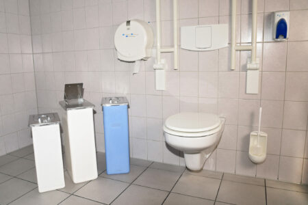 Ein öffentliches Badezimmer mit hellgrauen Fliesen an Boden und Wänden. Links stehen drei rechteckige Hygienebehälter: zwei in Weiß und einer in Blau, wobei einer der weißen Behälter eine geöffnete Klappe zeigt. Rechts im Bild befindet sich eine weiße Toilette mit geschlossenem Deckel neben der ein Toilettenbürstenhalter an der Wand befestigt ist. Über der Toilette sind Rohre und eine Notfallalarmkordel sichtbar. An der Wand sind zudem ein weißer Papierhandtuchspender und ein blauer Lufterfrischer installiert.