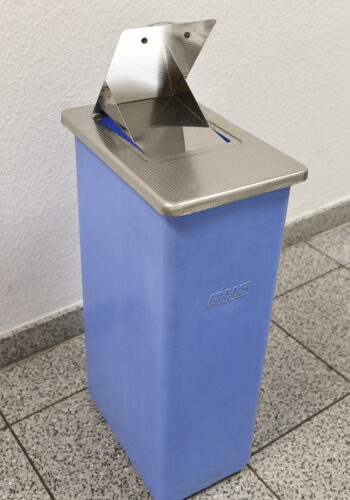 Ein schlanker, blauer Hygienebehälter steht auf einem grau gesprenkelten Fliesenboden gegen eine weiße Wand. Der Behälter hat eine glänzende, silberne Oberseite mit einer runden Öffnung, die durch eine Klappe aus Edelstahl mit scharfen Winkeln geschützt ist, die zur Benutzung offen steht. Auf der Vorderseite des Behälters ist das Logo 'BHS' in einem unaufdringlichen Grauton zu sehen