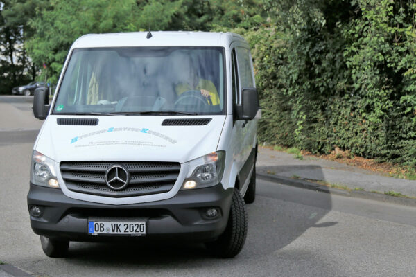 Ein weißer Mercedes Transporter fährt auf einer städtischen Straße, begleitet von üppigem Grün am Straßenrand. Das Fahrzeug, trägt Werbung für Hygiene-Service-Kegenhof, das Hygienehelfer, Aircare-Geräte und Hygieneartikel anbietet. Im Fahrzeug ist eine Person in gelber Kleidung am Steuer sichtbar, die konzentriert auf die Straße blickt.